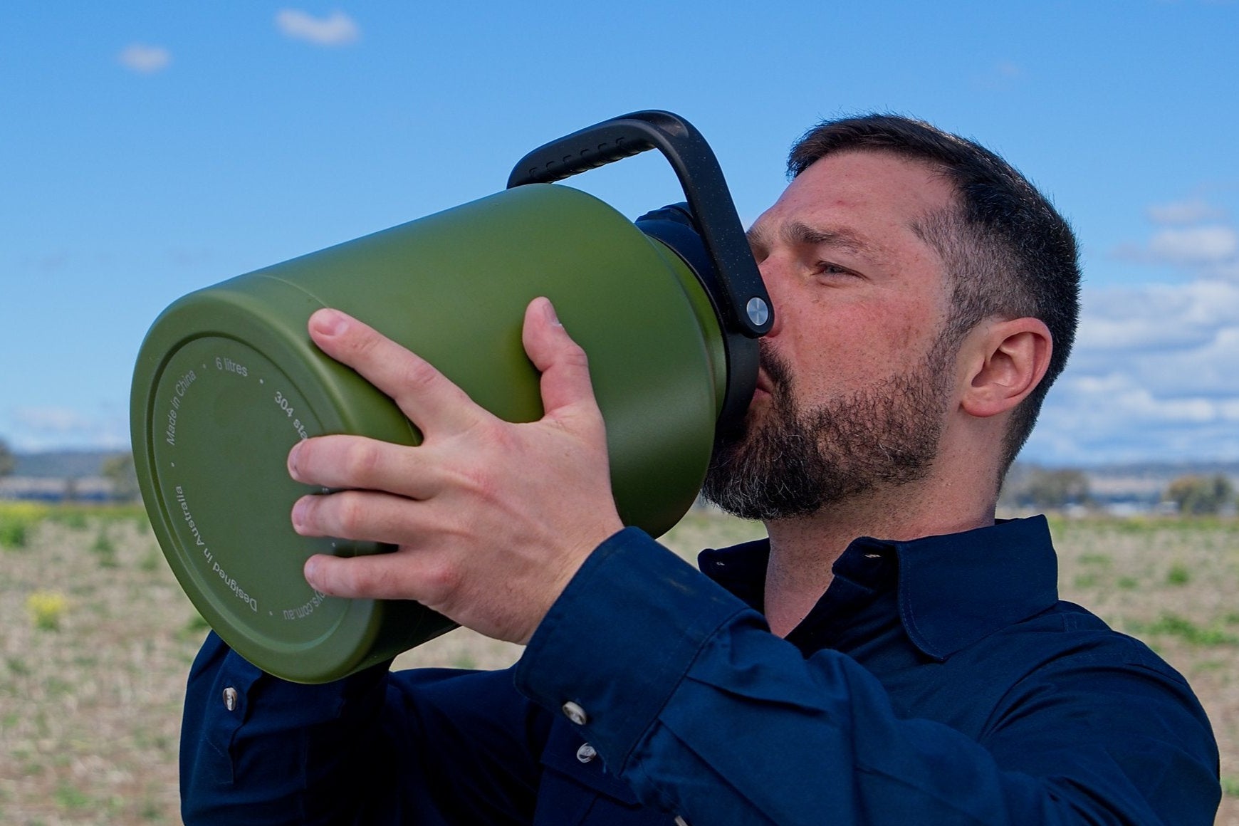 Man drinking from a green thermos in an outdoor setting with a clear sky.