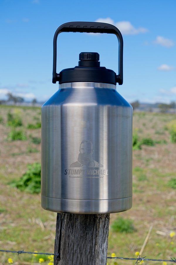 Stainless steel water bottle with handle on a wooden post against a rural landscape