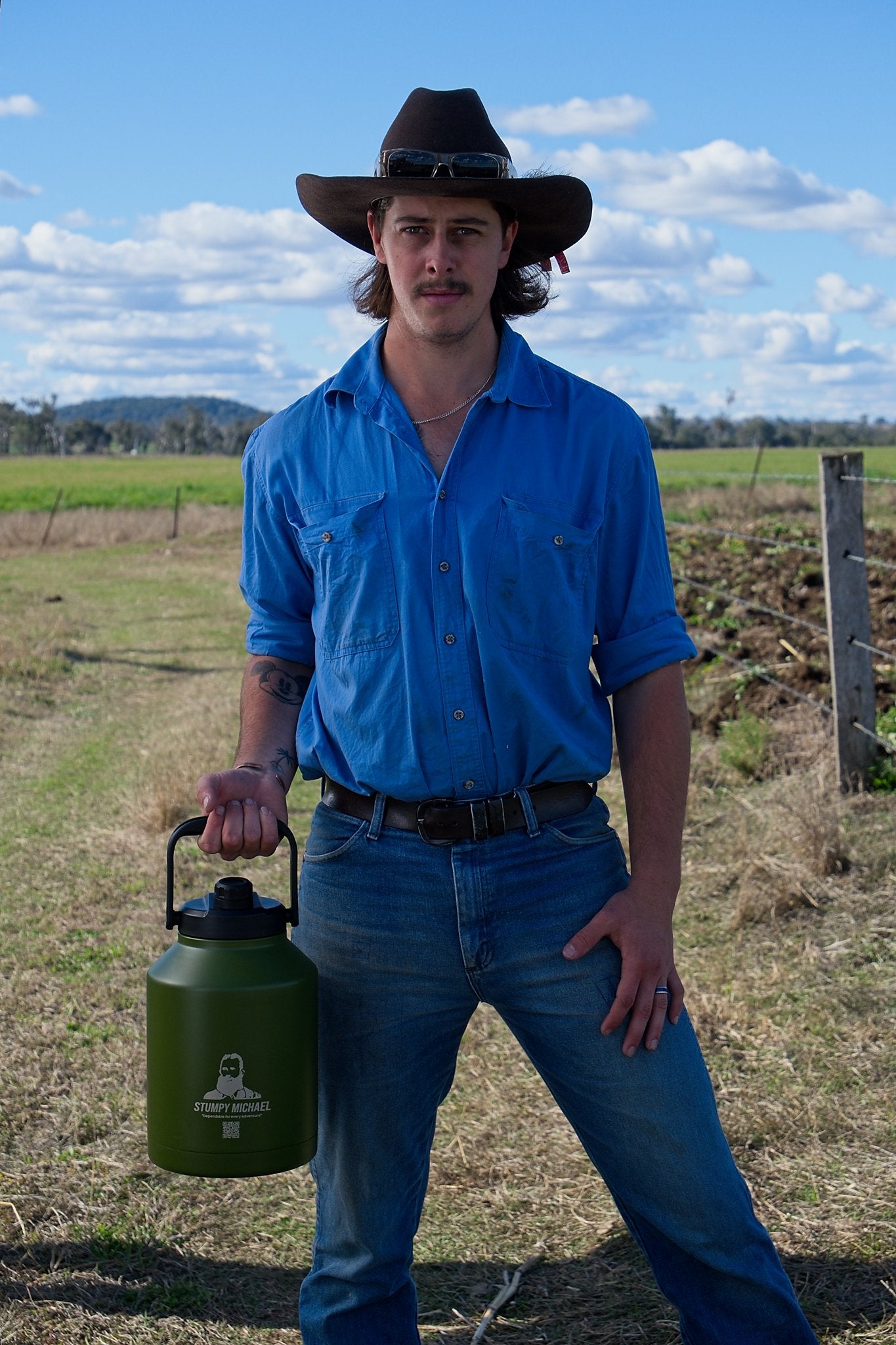 Man in a blue shirt and cowboy hat holding a green container in an open field.