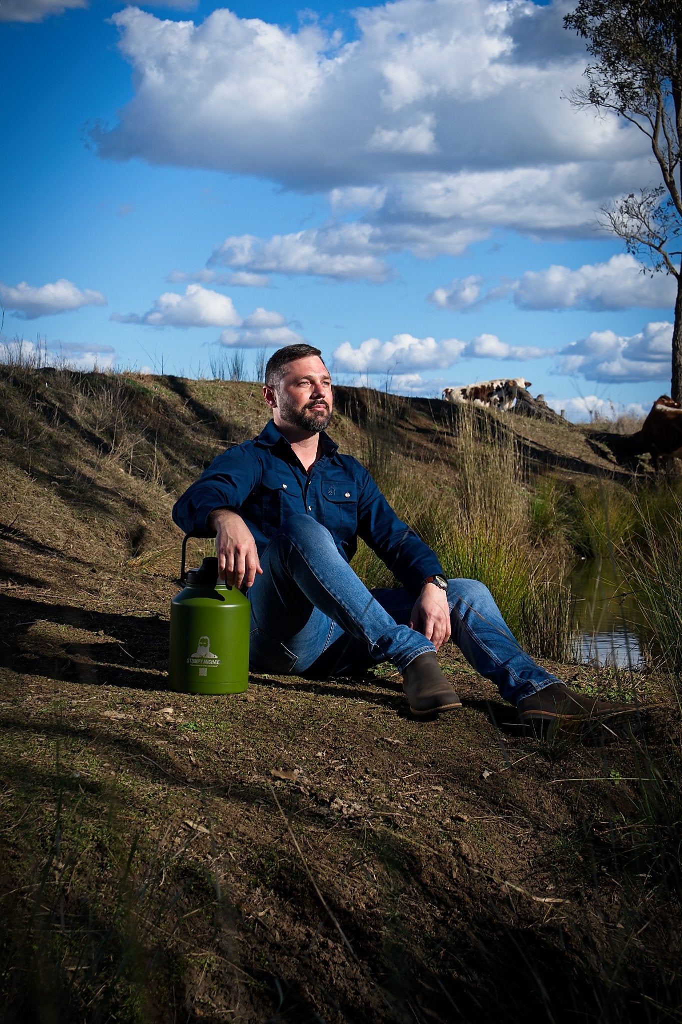 Man sitting on a hillside with a green container, under a blue sky with clouds.