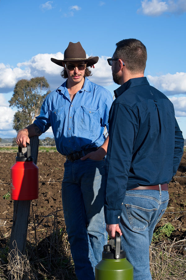 Two men standing in a field, one holding a red tool and the other a green tool, with a blue sky and clouds in the background.